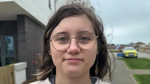 Patricia Czapka, is pictured on a road side with a police car behind her. She is wearing glasses and has brown hair. 