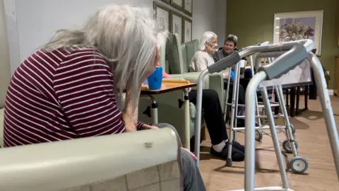 A woman with shoulder length grey hair with her head in her hands sitting in the foreground and a walking frame in front of her and two other women sitting in background