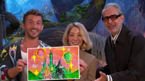 BBFC Group shot of Jonathan Bailey, Michelle Yeoh and Jeff Goldblum smiling as they hold up a copy of Maidah's artwork. They stand in front of artifical trees uses as backdrop props.