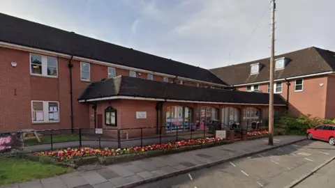 The front entrance to the Lichfield District Council office building. It is a two storey orange brick building that has a footpath with railing outside its frontage, which is lined with a flower bed