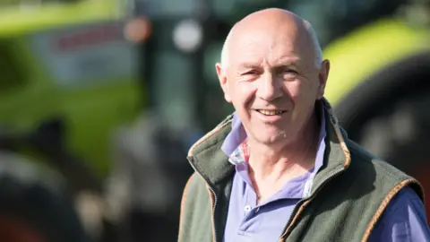 Countryside Regeneration Trust Farmer Tim Scott. He is bald with short grey hair at the sides. He is sun-tanned and is wearing a green sleeveless warmer and a blue top. The background, which shows farm machinery, is out of focus.