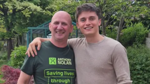 Two men stand smiling at the camera. The man on the left has a bald head and wears a green t-shirt saying ANTHONY NOLAN - SAVING LIVES. The younger man on the right, wearing a light coloured top, has his arm round the shoulder of the other man. They stand in a garden full of trees.