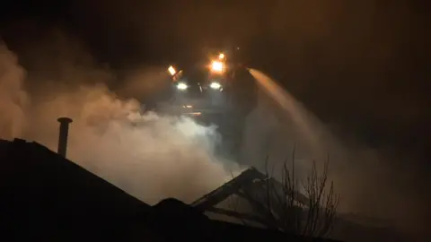 An aerial ladder with fire crew on board sprays water over a smoking building at night.