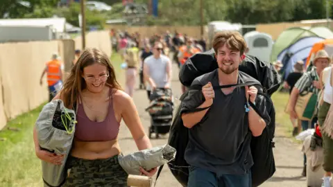 Sarah Louise Bennett A young man and young woman are among people carrying camping equipment on a path on the festival site.