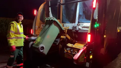 A man in high-vis smiles at camera whilst a bin is being taken up into the back of the lorry.
