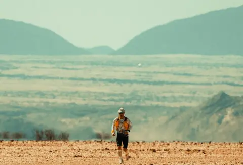 Jenny Scott Communications Jon Shield is seen from head to toe running - a small part of the image near the bottom in the centre. A mountain landscape is in the distance. 