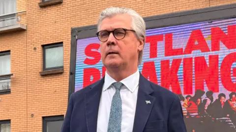 PA Media Malcolm Offord, who has grey hair and glasses, wearing a business suit while standing in front of a building and a digital billboard 