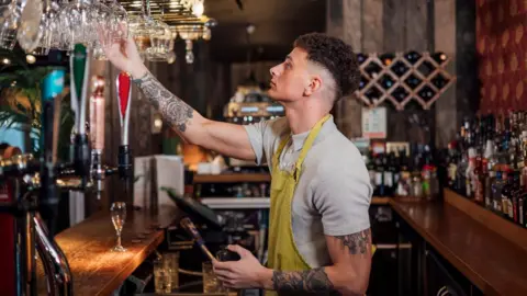A bar staff member wearing a grey t-shirt and a yellow apron stands behind the bar holding a glass in his hand. 