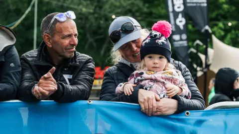Red On Sports A young girl with a pink bobble hat and blonde hair watches a cycling race, she is being held by a woman with blonde hair and a blue hat with glasses resting on it. A man with his sunglasses tipped back on his head with short greyish-black hair and a leather jacket is next to them.
