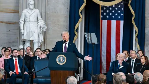 US President Donald Trump speaks after being sworn-in. He stands at a podium with the presidential seal and is flanked by a seated crowd that includes Vice-President JD Vance and former President Joe Biden. A US flag hangs behind him. 