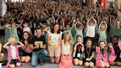 A hall full of children with their hands in the air cheering with three adults at the centre front