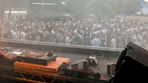 A set of DJ decks in the foreground with a crowd of people in the background, at a music festival. There's trees in the background behind the crowd. The crowd themselves are dancing, all are wearing various items of summer gear like shorts, T-shirts and bucket hats.