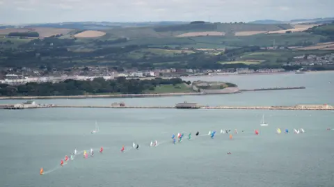 PA View of Portland Harbour with Weymouth in the background. A long line of small boats with different coloured sails runs from right to left across the picture.