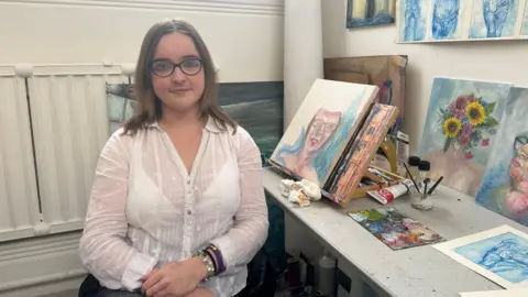 Zoe Dennis/BBC Poppy sitting down with her art work series in the background