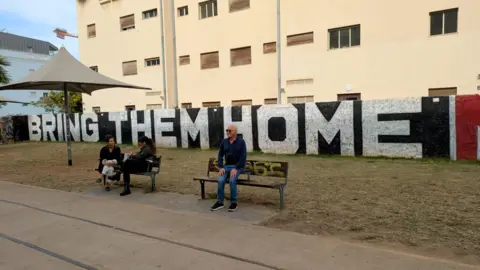 BBC People sitting in front of a mural displaying words Bring Them Home in Tel Aviv