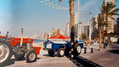 Dave Harrison Two tractors parked on the promenade in Benidorm. The weather is sunny and hotels, palm trees, sand and sea are visible in the background.