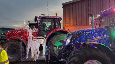 A side-on image of two tractors decked in multi-coloured Christmas lights, parked outside a brown building. The left tractor is red and the right is blue. In front of the red tractor is two reindeer statues made of white Christmas lights.