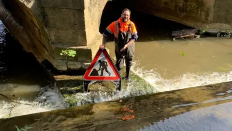 A council worker wearing orange high vis and wellington boots is standing in a river holding a road sign. The river is flowing behind him and there is a rusty trolley in the water.