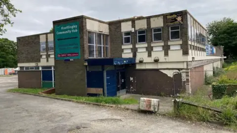 A tired-looking two-storey 1960s building, surrounded by a narrow grass verge and tarmac. A silver fence, which is blocking off access to a path down one side of the building, sits to the side of the picture.