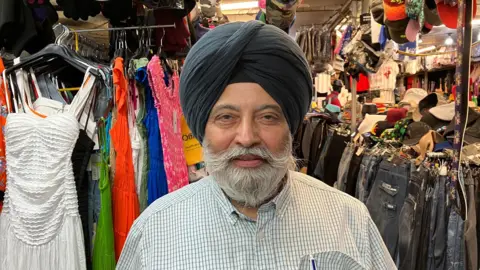 Sam Read/BBC A picture of a man with a turban on, who also has a full beard and a moustache. He is stood in front of rows and rows of clothes that he sells in his market stall.