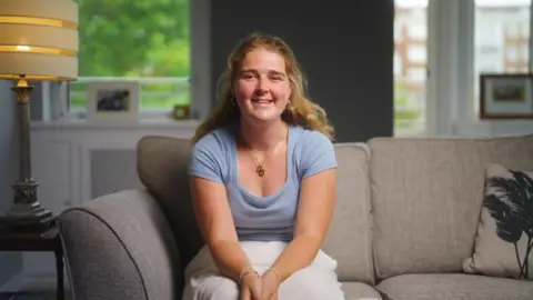 Children with Cancer UK Libby sitting on a sofa, smiling at the camera, wearing a pale blue t-shirt and white skirt.