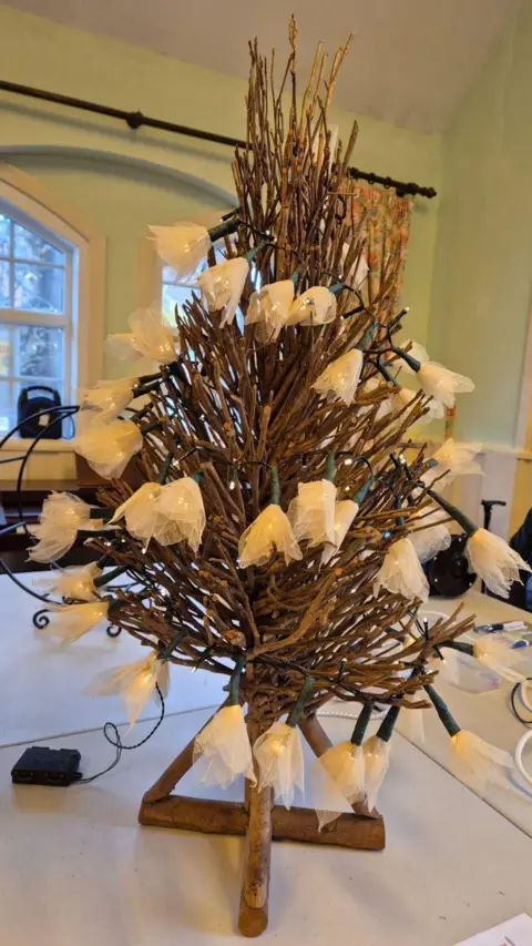 The Studley Repair Cafe A small Christmas tree with brown branches sits on a table, covered in fairy lights with white leaves surrounding them.