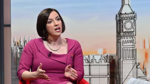 Bridget Phillipson, a woman with dark hair in a bob, wearing a fuchsia pink outfit in front of a stylised graphic of the Houses of Parliament on the set of Sunday with Laura Kuenssberg