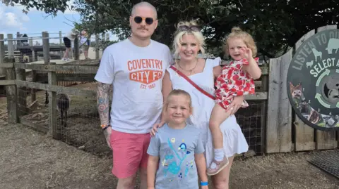 Hannah Deez Hannah's family of four stand outdoors on a gravel path near a wooden animal enclosure. She is holding a small child in patterned clothing, while another child stands at the front. A large sign and trees form part of the background.