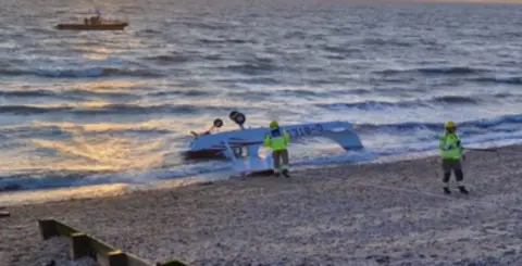 A light aircraft sits upside down in shallow water by the edge of a beach. Two people in high-vis jackets stand nearby
