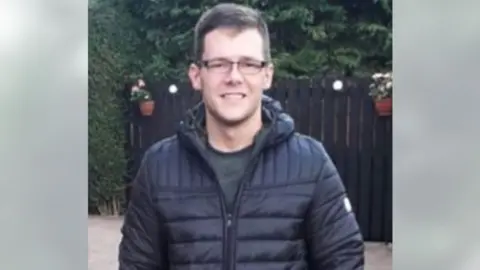 Police Scotland A smiling man with short brown hair photographed in a garden. He is wearing brown glasses and a blue jacket.