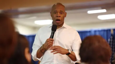 Eric Adams speaking into a microphone. he is wearing a white shirt with rolled up sleeves.