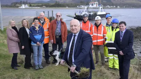 The Highland Council A group of Highland Councillors are standing in front of a slipway next to Loch Linnhe. A ferry can be seen in the background. One of the councillors is holding a shovel with a newly cut piece of turf on it.
