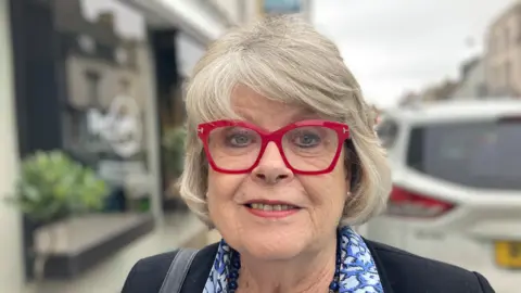 A woman with red glasses looks directly at the camera, she has grey bobbed hair. She is wearing a jacket and a blue patterned shirt. She has a handbag strap over her shoulder and is wearing a necklace and earrings.