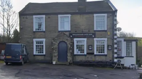 Google Exterior of the Roggeram Gate pub in the Briercliffe area of Burnley. It is a stone-built, double-fronted pub, with a hardstanding area in front. 