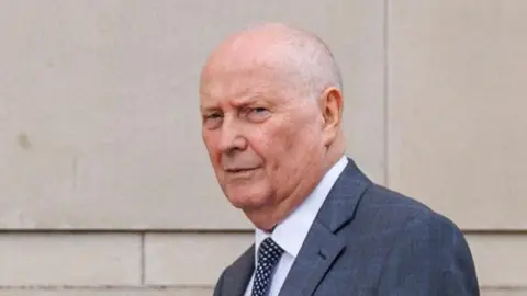 Frank Cushnahan, pictured outside Belfast Crown Court, has thin grey hair and is wearing a grey suit jacket, white shirt and a navy polka dot tie.