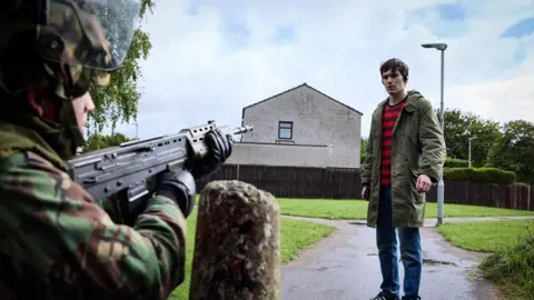 Keeper Pictures and Cyprus Avenue Films A crouched solider point his gun at a young man in a green parka coat. Behind him is a terrace house, a lamppost, and a path that forks through a green. 
