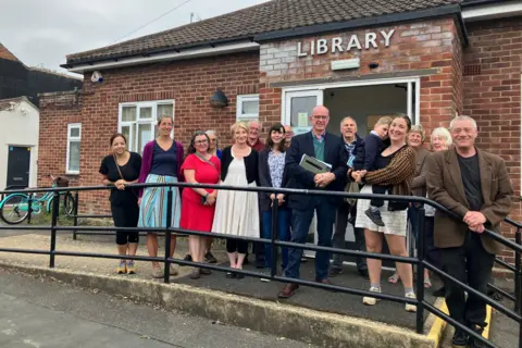 Shariqua Ahmed/BBC A group of people stand in front of a small red bricked building with the word 'library' written on its front. There are 15 people standing in front of a railing including a woman with a small child