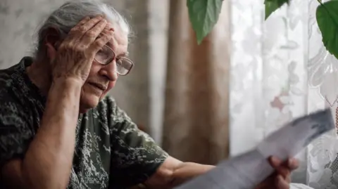 An elderly woman, wearing a green and whites, sits at a table and puts her hand to her head while reading an energy bill