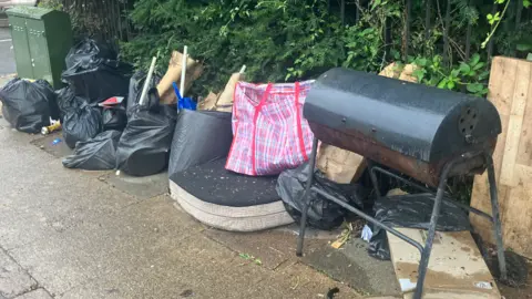 Nadia Lincoln/ LDRS Rubbish dumped on Semilong Road, Northampton. It includes a black barbeque on legs, black plastic bin bags with cardboard sticking out, and a seat cushion.