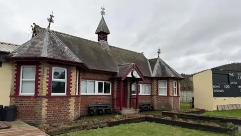 An old Victorian era cricket club with a pointed part sticking out the top of the roof. The window, the door and its trim are painted a maroon colour. There is a analogue clock above the entrance way.