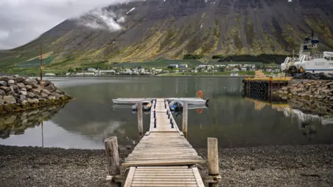 Universal Images Group via Getty Images A lake with a wooden jetty built on gravel land going out into water. There are small islands of rocks either side. In the distance is a mountain with low cloud descending.
