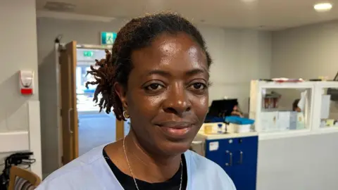 Folake looking to the right of the camera. She is wearing her blue care home scrubs with a thin gold chain and a black layer underneath. Her dark hair is tied back. A care home reception desk can be seen to the right hand side, with an open door behind her.