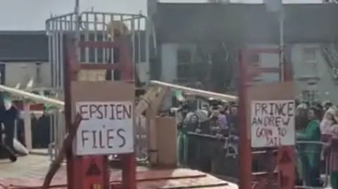 Social Media A screenshot of the back of a tractor trailer. There is a cage with a person in it. There are also signs reading 'Epstien files' and 'Prince Andrew goin to jail'.
