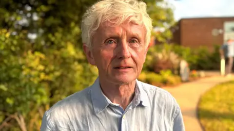 Seamus O'Brien pictured outside with trees and a path behind him. He is wearing a light blue buttoned shirt and has wavy white short hair.
