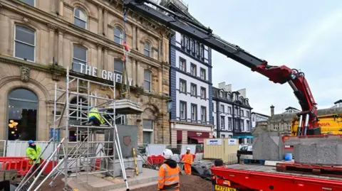 A large monument - a drinking fountain dating from the 1860s - being installed on a stepped set of foundations. A giant crane is lowering a stone cap onto its rectangular base. A workman is kneeling on scaffolding to supervise the join as others in high viz uniforms look on. The work is going on in front of a sandstone building with the appearance of a hotel and a sign that reads The Griffin.