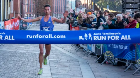 Run For All Runner in blue with green trainers lifting his arms as he's about to cross the finish line 