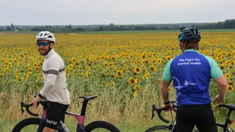 Supplied Two men are bikes pause near a sunflower field. One cyclist's jersey reads: "We fight for mental health" 