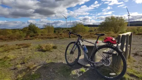 A bike sitting next to a wooden chair and table, looking over greenspace with wind farms in the background. A red cyclist's helmet is sitting there too. 
