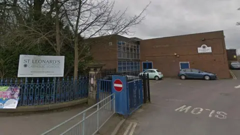 Streetview of a school entrance with a sign saying St Leonard's Catholic School next to it. Beyond the blue gate can be seen a two-storey brick building with large windows and several parked cars.