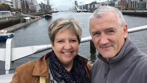 The Stroke Association A woman in a brown coat and black scarf stands next to a man in a grey zipped up fleece on a bridge overlooking a river. Another bridge can be seen in the distance. 